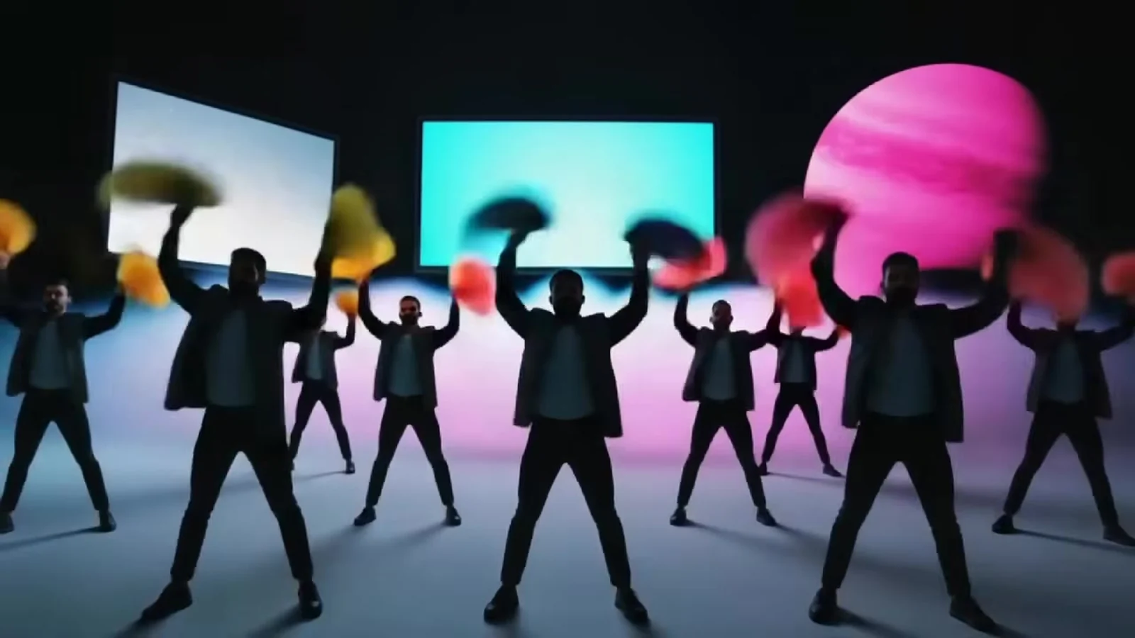Fotograma del videoclip de «Discutiendo con la IA»: siluetas negras con abanicos de colores en alto delante de pantallas turquesa y un planeta rosa con franjas
