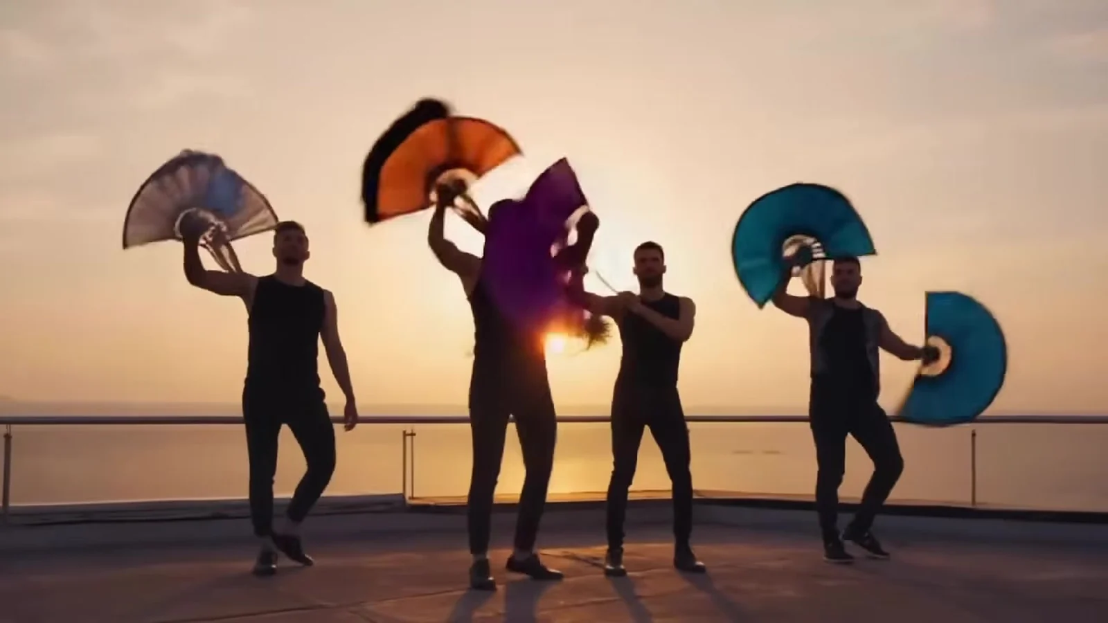 Fotograma del videoclip de «Discutiendo con la IA»: cuatro bailarines a contraluz junto al mar alzan abanicos rojo, azul, morado y naranja mientras se pone el sol