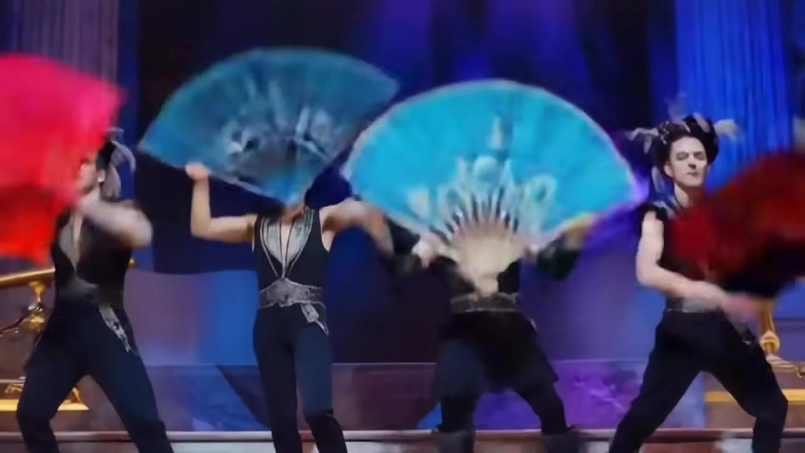 Fotograma del videoclip de «Discutiendo con la IA»: abanicos de tela turquesa y plumas rojas se abren en un escenario azul y tapan a los bailarines