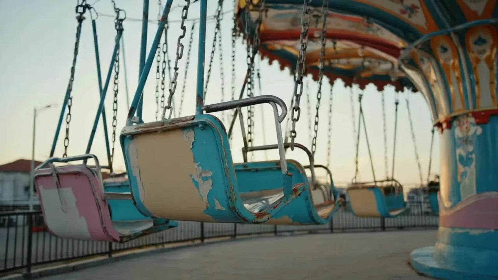 Fotograma del videoclip de «Boys from the Village»: las sillas voladoras de una feria, vacías y quietas, con los asientos azul celeste y rosa colgados de cadenas y la pintura descascarillada