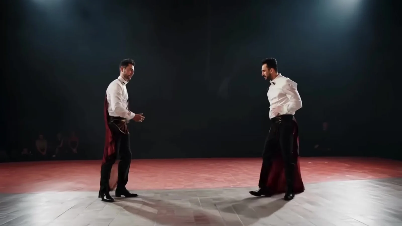 Fotograma del videoclip de «Carta de recomendación»: dos bailarines con camisa blanca y falda larga, frente a frente sobre una tarima iluminada en un escenario oscuro