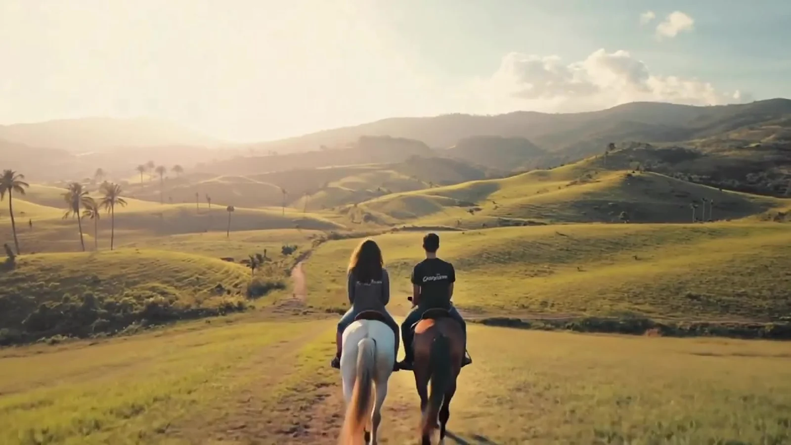Fotograma del videoclip de «Que isso»: dos jinetes de espaldas, uno en un caballo blanco y otro en uno castaño, avanzan juntos por un camino de tierra en un valle verde con palmeras y colinas al amanecer