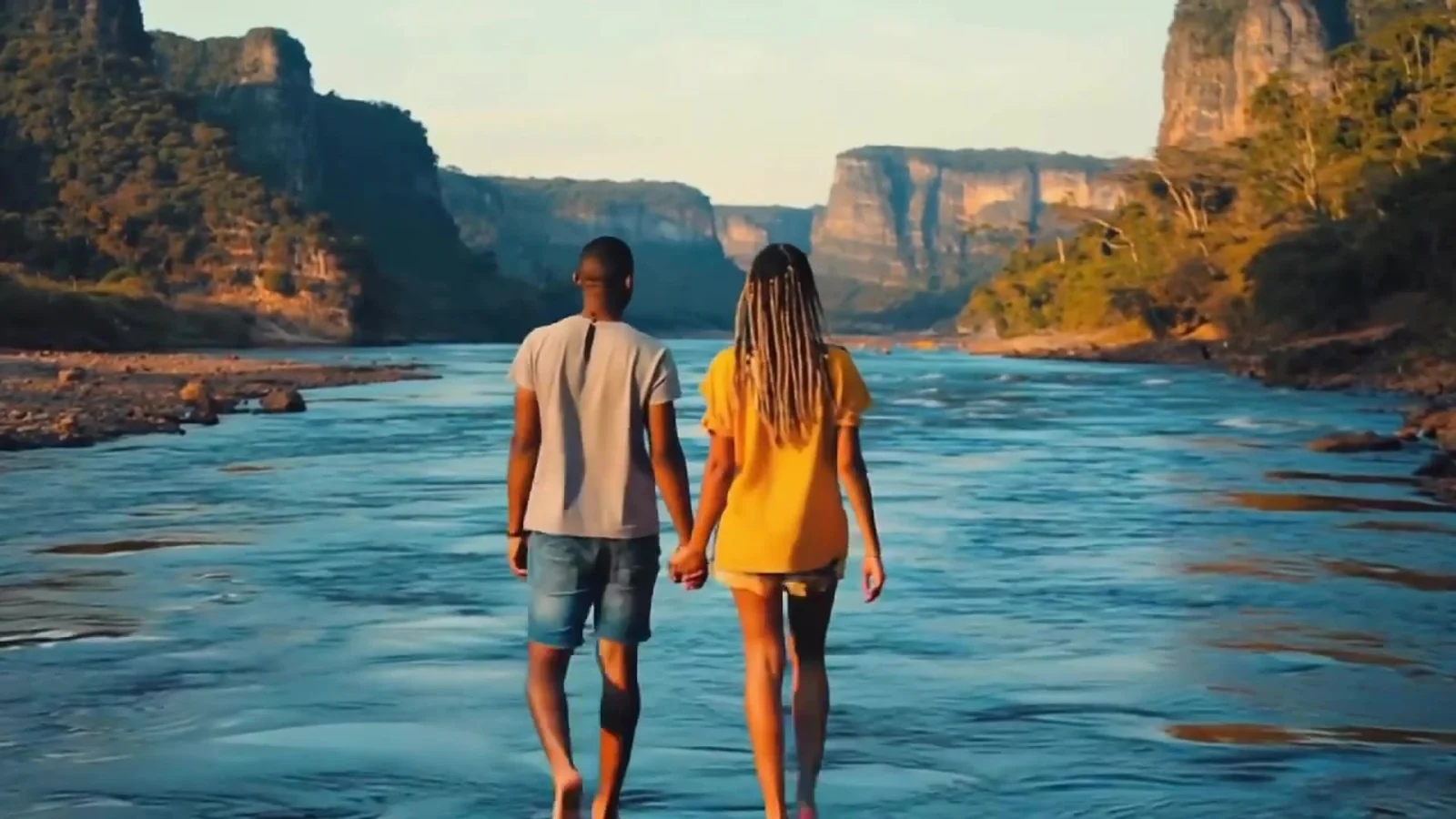 Fotograma del videoclip de «Que isso»: una pareja de espaldas camina de la mano por un río de aguas azules entre altas paredes de roca rojiza