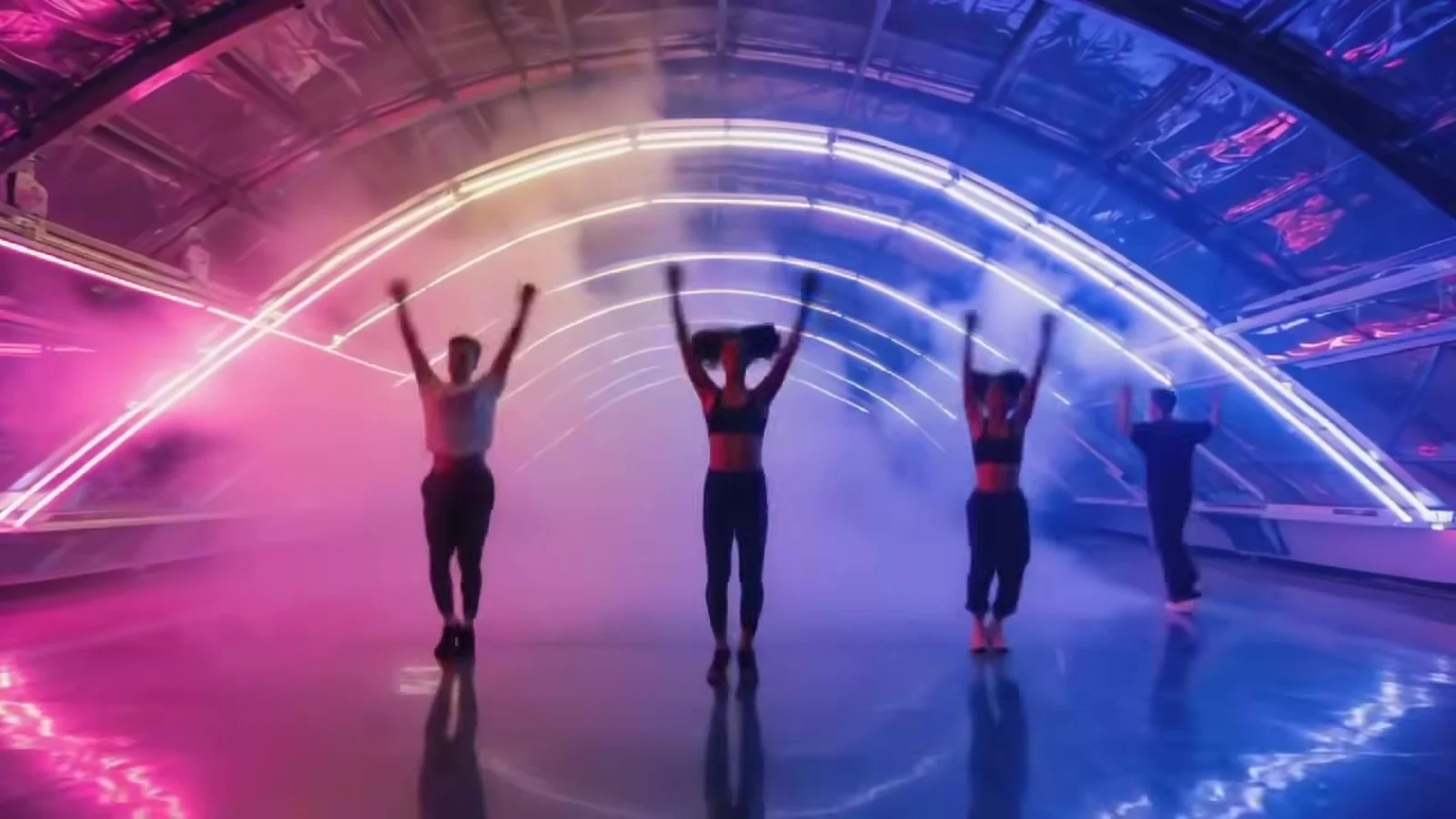 Fotograma del videoclip de «Zee-Zee»: cuatro bailarines de espaldas levantan los brazos bajo una bóveda de cristal cruzada por arcos de neón rosa y azul, con niebla sobre un suelo brillante