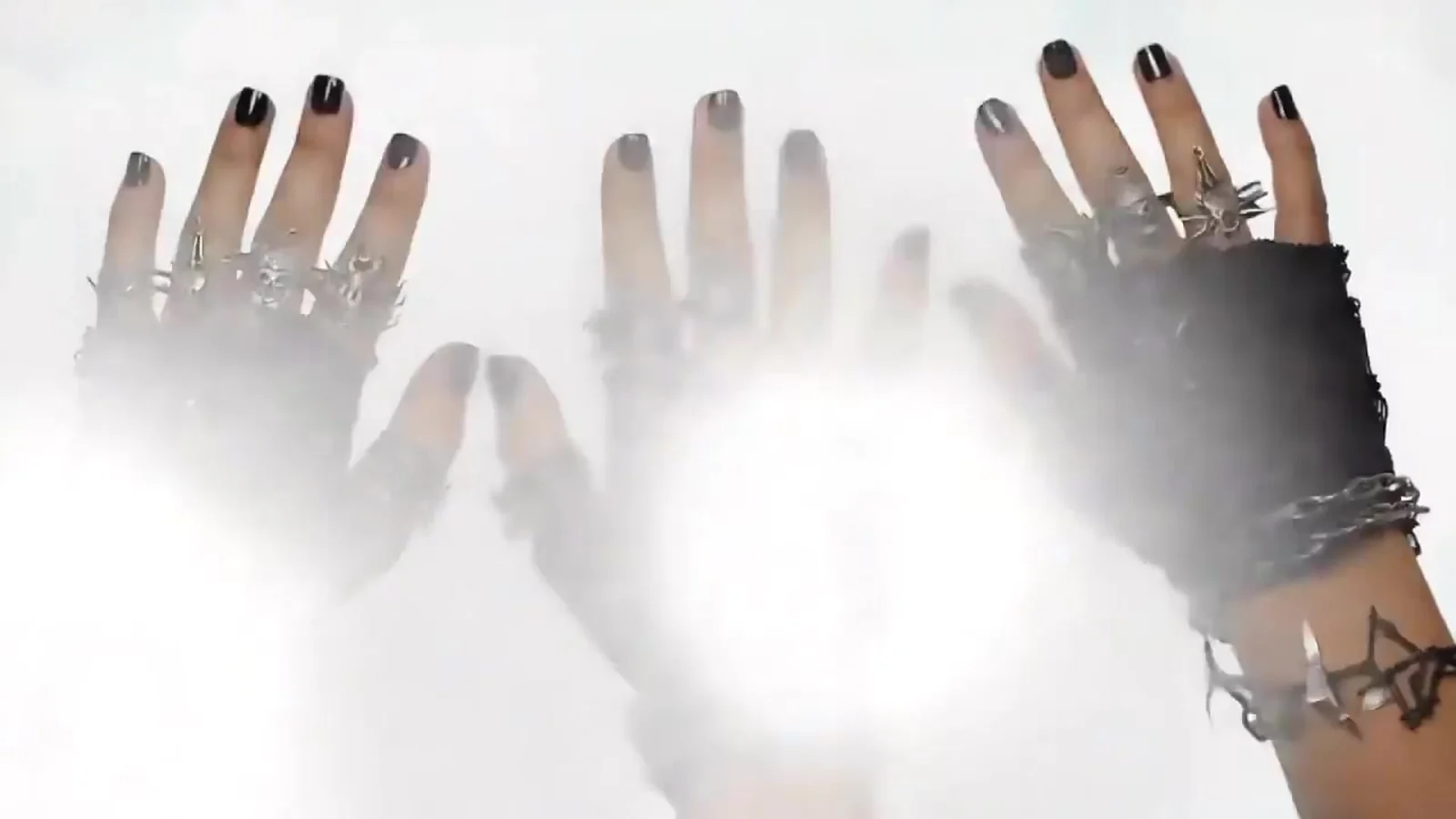 Fotograma del videoclip de «Beats Drop Time Stops»: varias manos con uñas negras, anillos de plata y pulseras de cadena se estiran hacia una luz blanca muy intensa