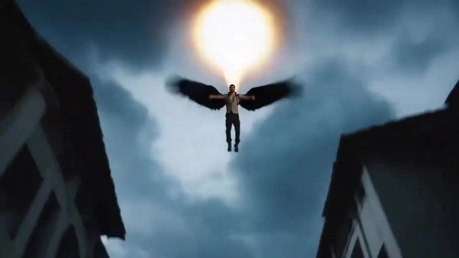 Fotograma del videoclip de «Fly Away»: una figura pequeña con alas negras abiertas suspendida en el cielo nublado, de espaldas a un sol blanco, entre dos tejados oscuros