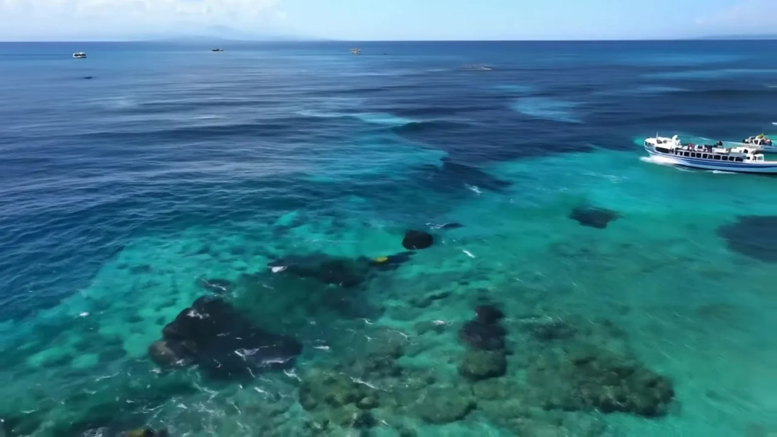 Fotograma del videoclip de «El canario que miente»: un mar turquesa con manchas oscuras de roca visto desde el aire y un barco blanco que deja una estela