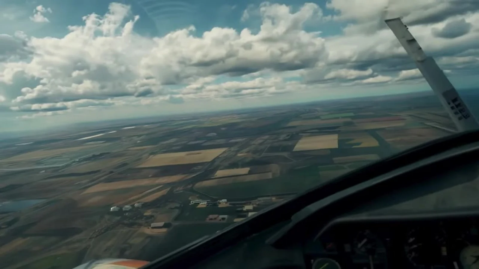Fotograma del videoclip de «El canario que miente»: desde la cabina, una llanura de parcelas verdes y amarillas bajo nubes de algodón durante el descenso