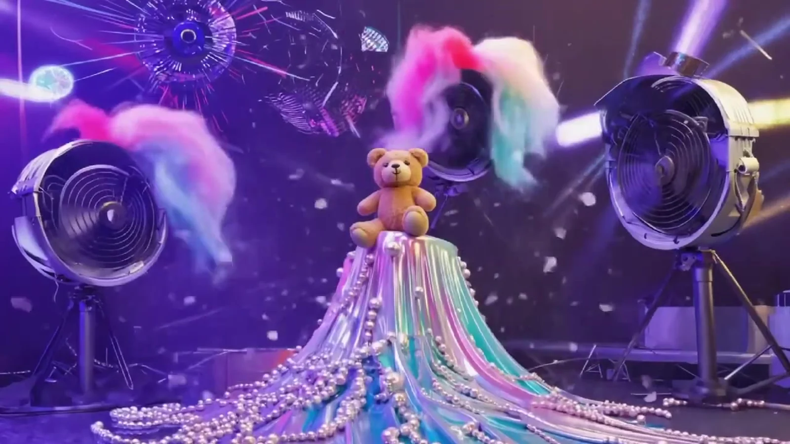Fotograma del videoclip de «Veneno»: un osito sentado en lo alto de una cascada de perlas mientras dos ventiladores le soplan plumas de colores