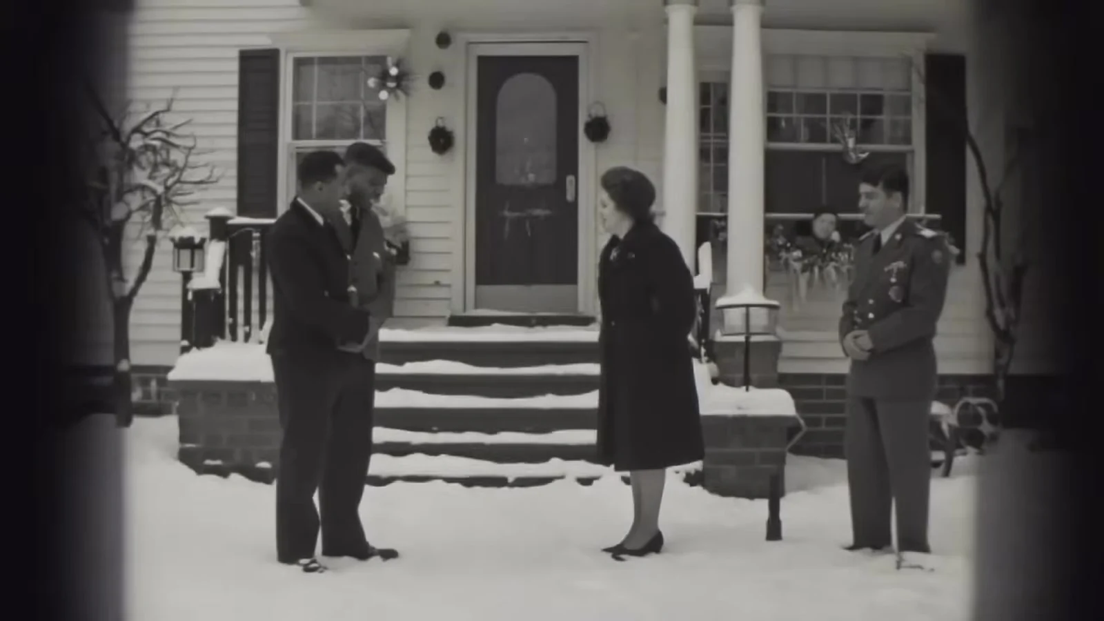 Fotograma en blanco y negro del videoclip de «Holy Stillness»: dos hombres con gorra de oficial y una mujer con sombrero, vistos de lejos ante los escalones nevados de una casa con columnas