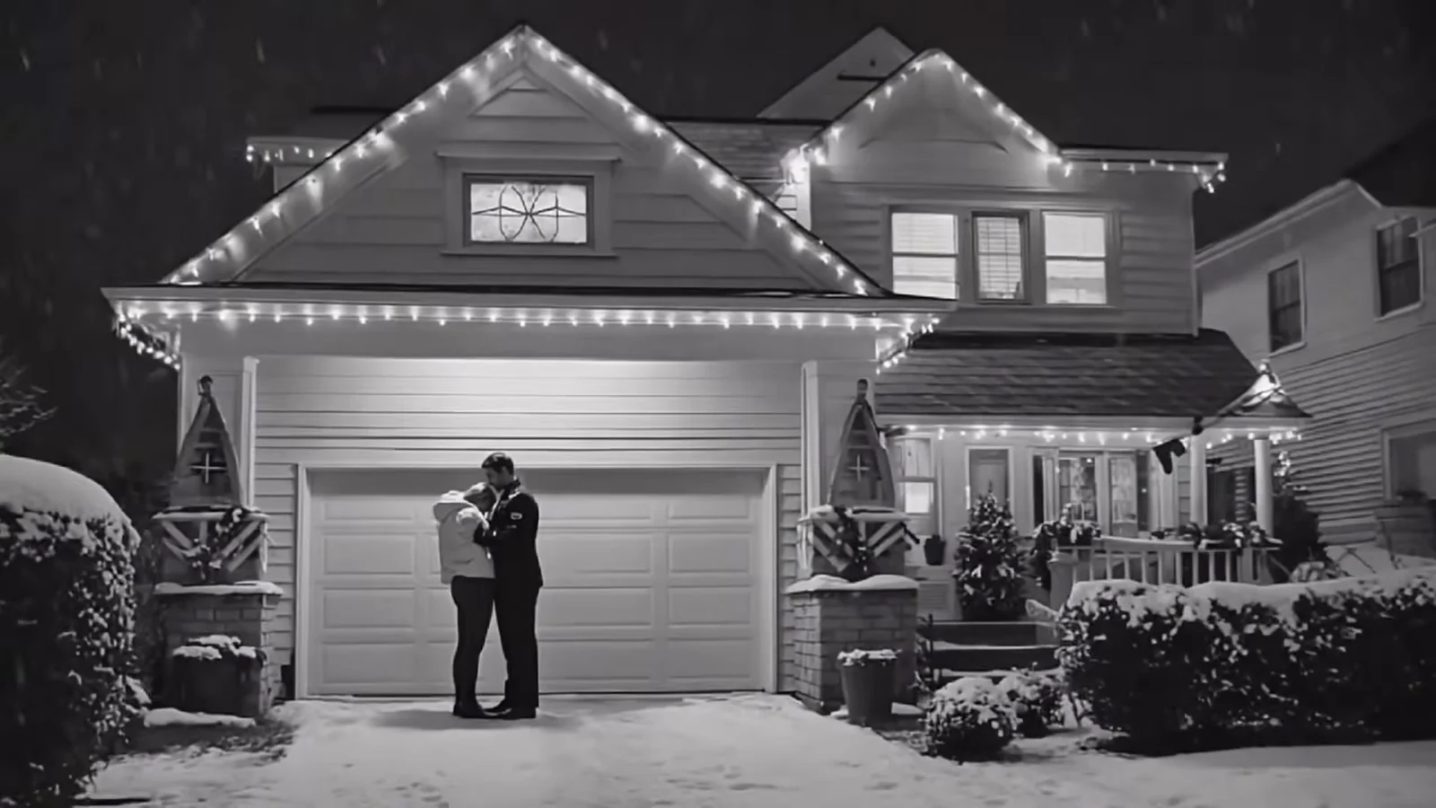 Fotograma en blanco y negro del videoclip de «Holy Stillness»: una casa nevada con el tejado bordeado de luces y una pareja diminuta abrazada frente al garaje
