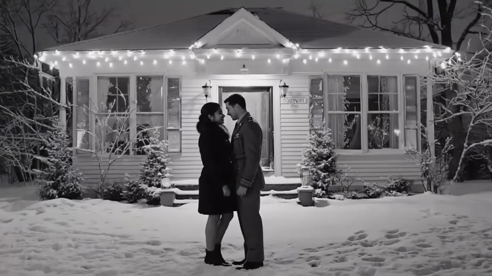 Fotograma en blanco y negro del videoclip de «Holy Stillness»: una pareja en la nieve delante de una casa con una guirnalda de luces en el porche