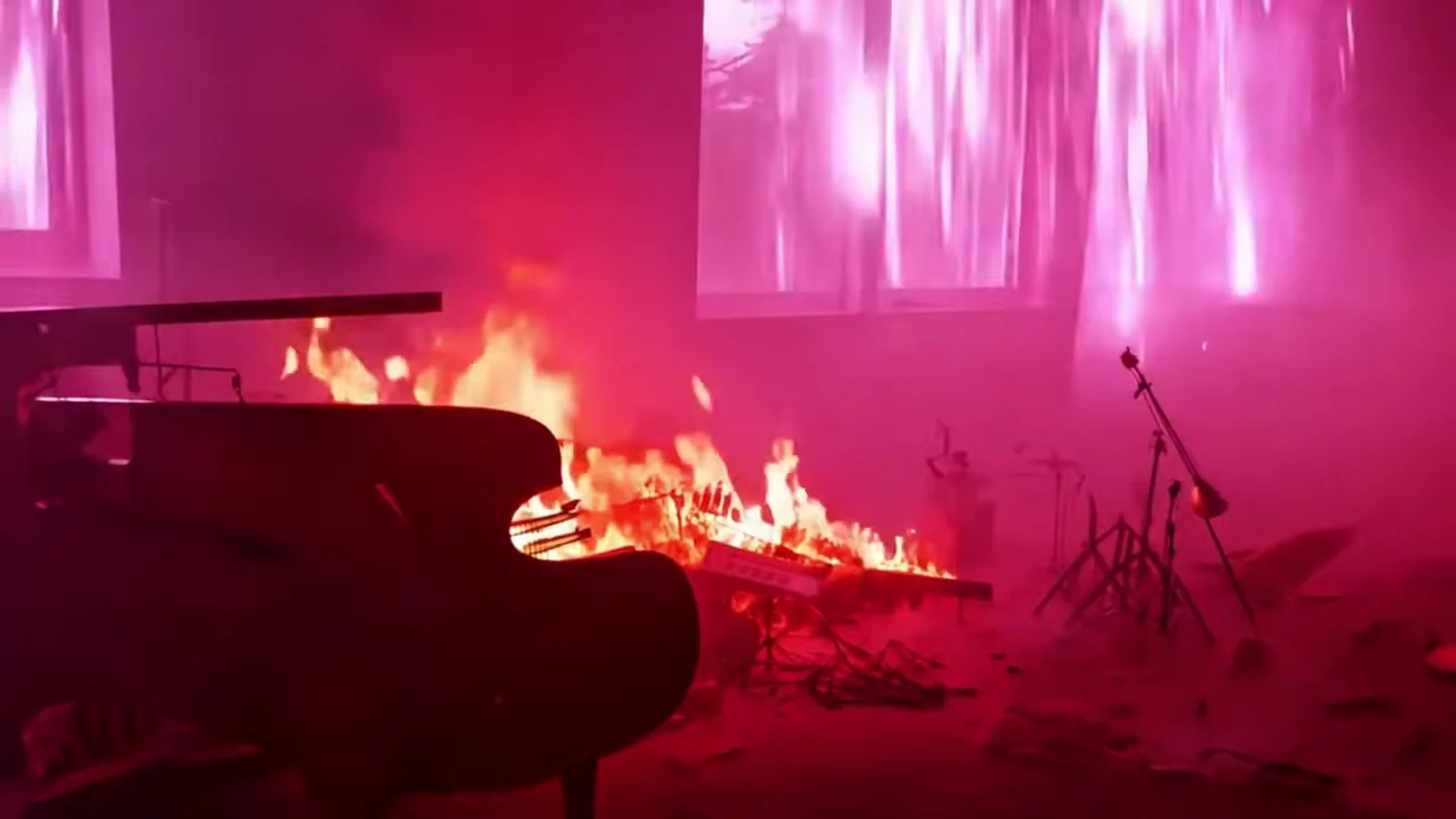 Fotograma del videoclip de «¡Vaya director!»: un piano de cola negro con llamas sobre la tapa en una habitación abandonada con ventanas de luz rosa y atriles de micrófono