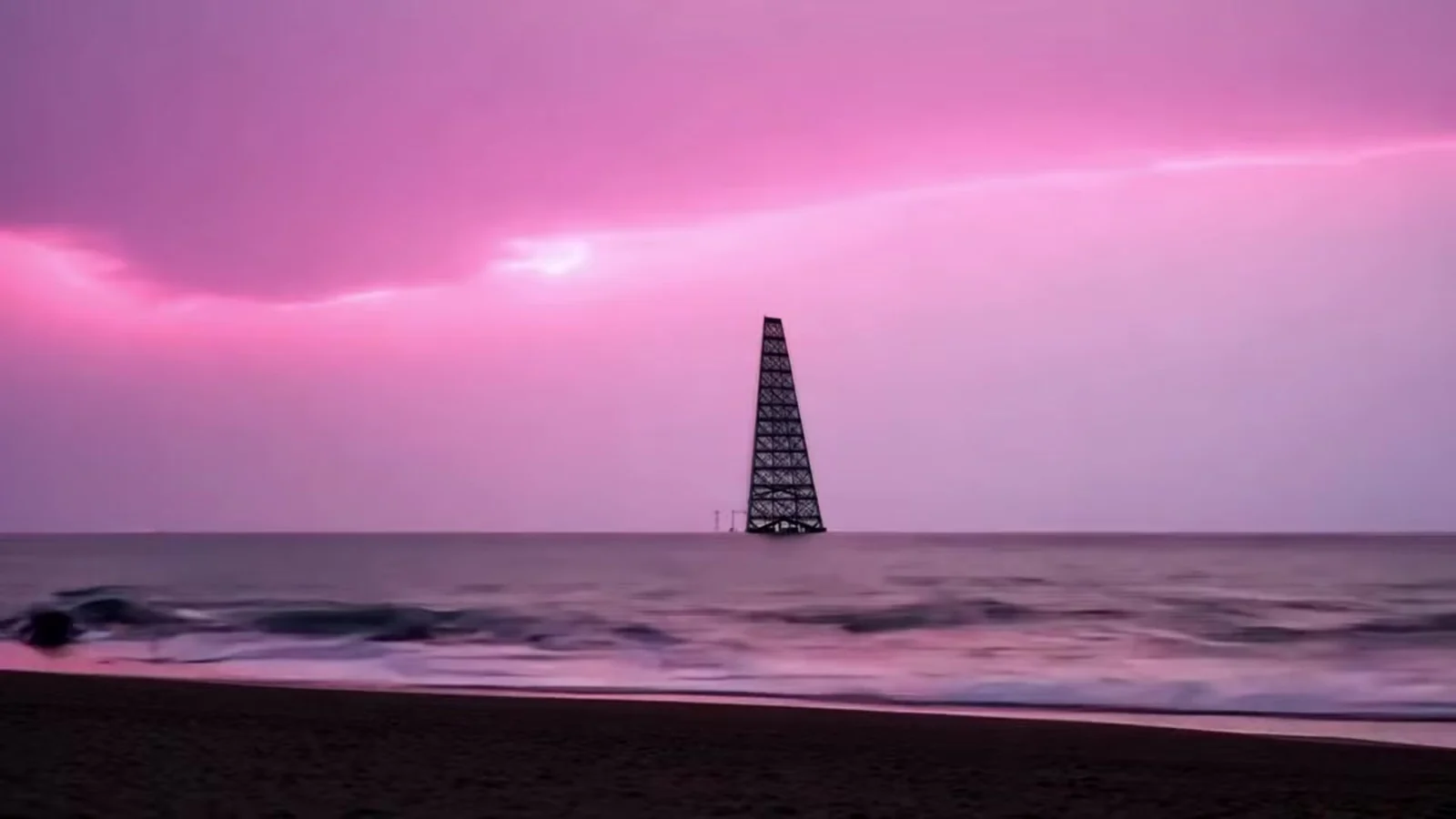 Fotograma del videoclip de «¡Vaya director!»: una torre de hierro solitaria en mitad de un mar tranquilo bajo un cielo completamente rosa, vista desde una playa oscura