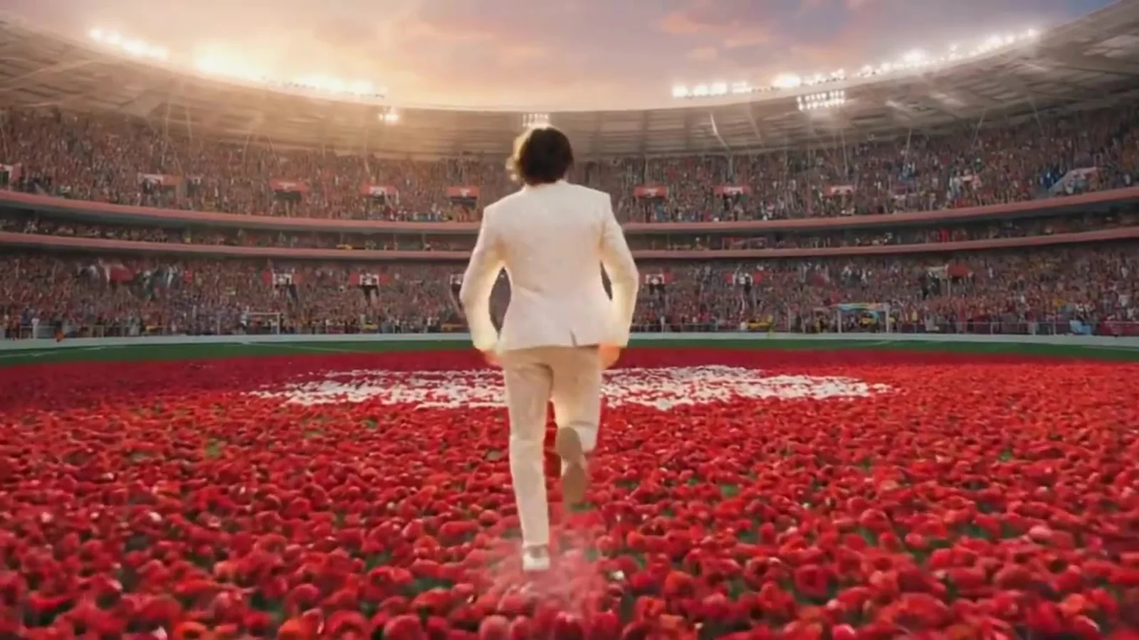 Fotograma del videoclip de «No me gusta el fútbol»: un hombre vestido de blanco camina de espaldas sobre una alfombra de pétalos rojos que cubre el césped, con la grada llena y los focos encendidos al atardecer