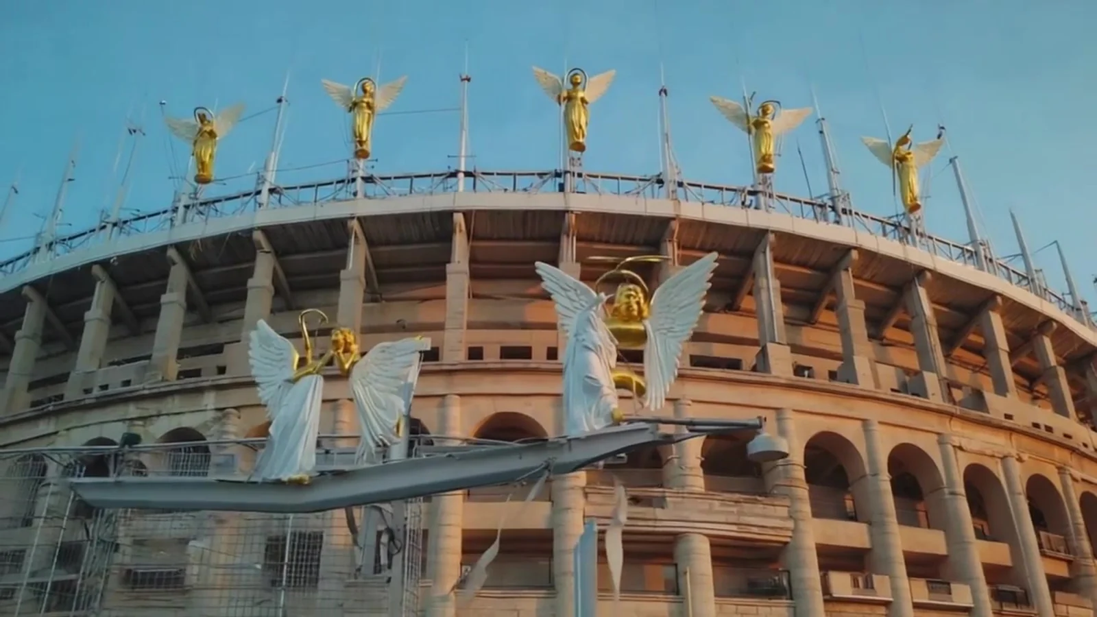 Fotograma del videoclip de «No me gusta el fútbol»: un estadio de arcos de piedra con una fila de ángeles dorados en lo alto del graderío y dos ángeles de alas blancas delante de los arcos