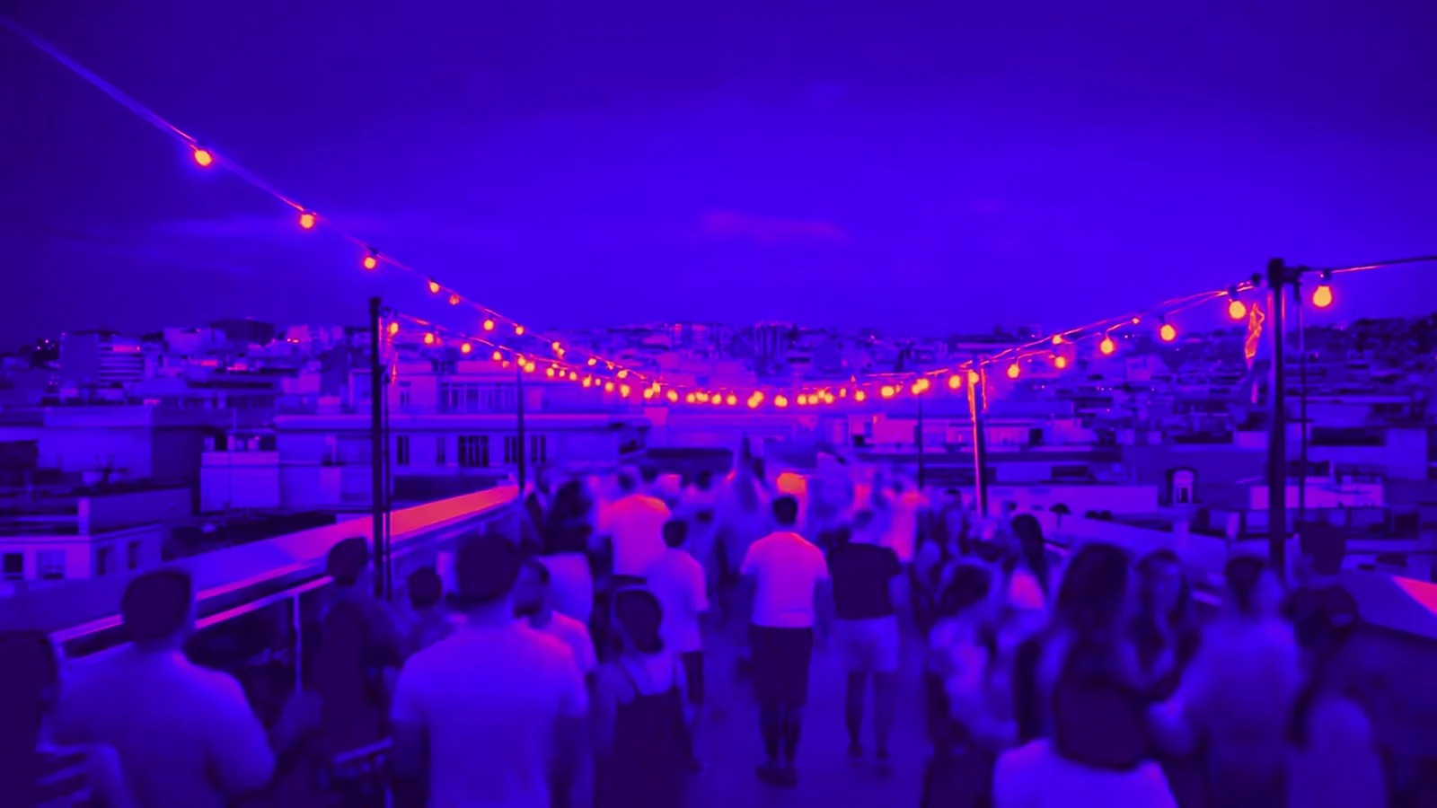 Fotograma del videoclip de «Barcelona»: la ciudad iluminada de noche vista desde lo alto, con haces de luz violeta y turquesa sobre los tejados