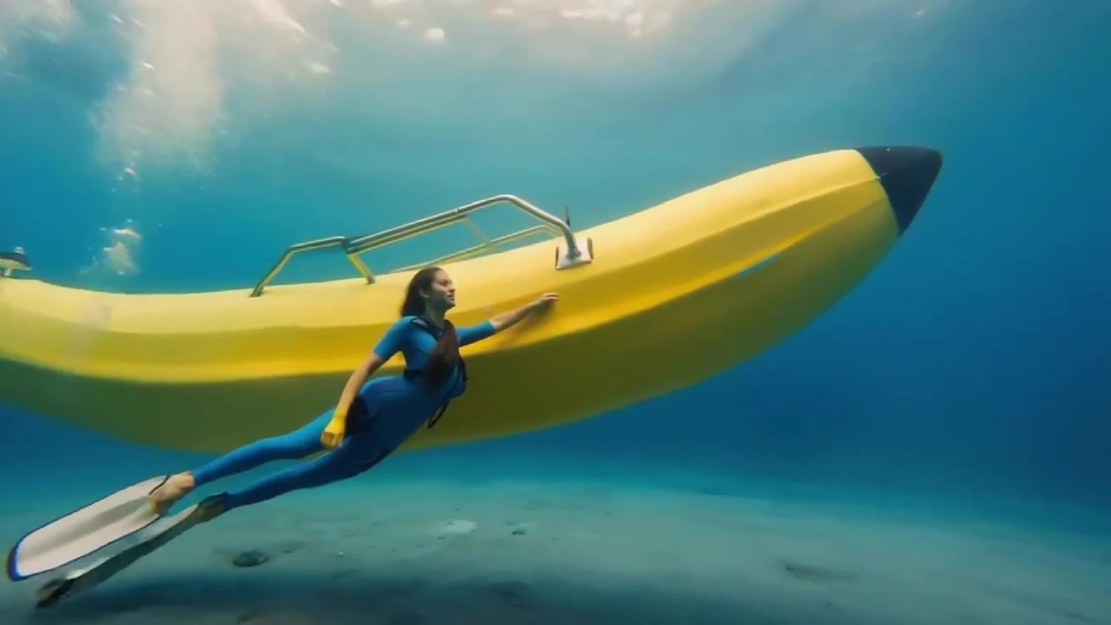 Fotograma del videoclip de «En Canarias no existen las sirenas»: un plátano hinchable amarillo gigante flota bajo el agua turquesa entre rayos de sol, con una figura con traje de buceo agarrada a él