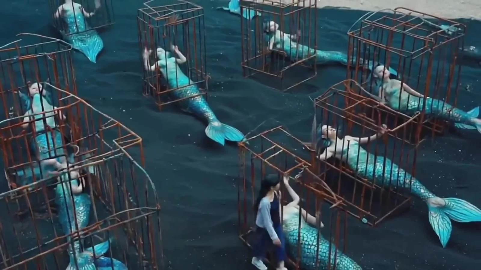 Fotograma del videoclip de «En Canarias no existen las sirenas»: vista desde arriba de un campo de jaulas en la arena del fondo del mar, con colas de sirena de color turquesa dentro de cada una