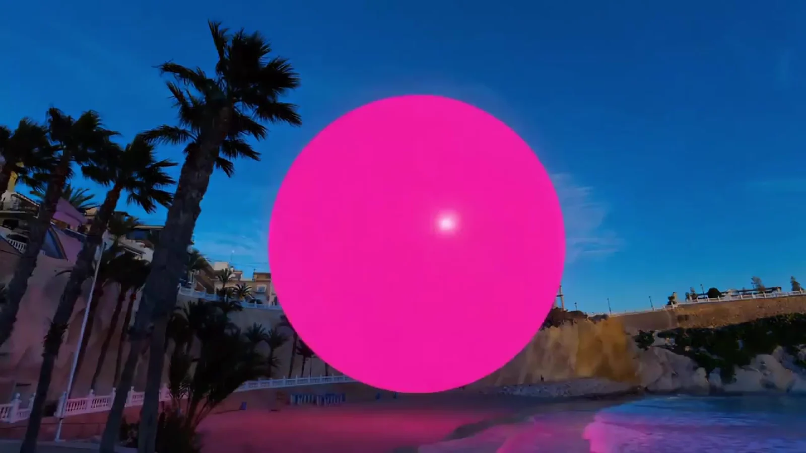 Fotograma del videoclip de «La playa del Mal Pas»: una esfera rosa enorme flota sobre una cala del Mediterráneo, detrás de los troncos y las hojas de unas palmeras, bajo un cielo azul
