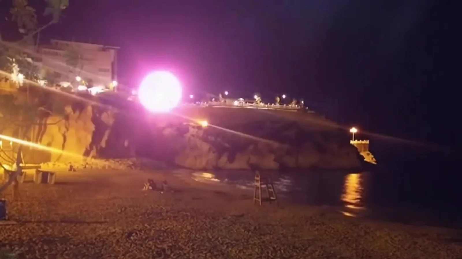 Fotograma del videoclip de «La playa del Mal Pas»: una luz rosa blanquecina brilla a ras de la orilla en una playa a oscuras, con el acantilado iluminado por farolas amarillas