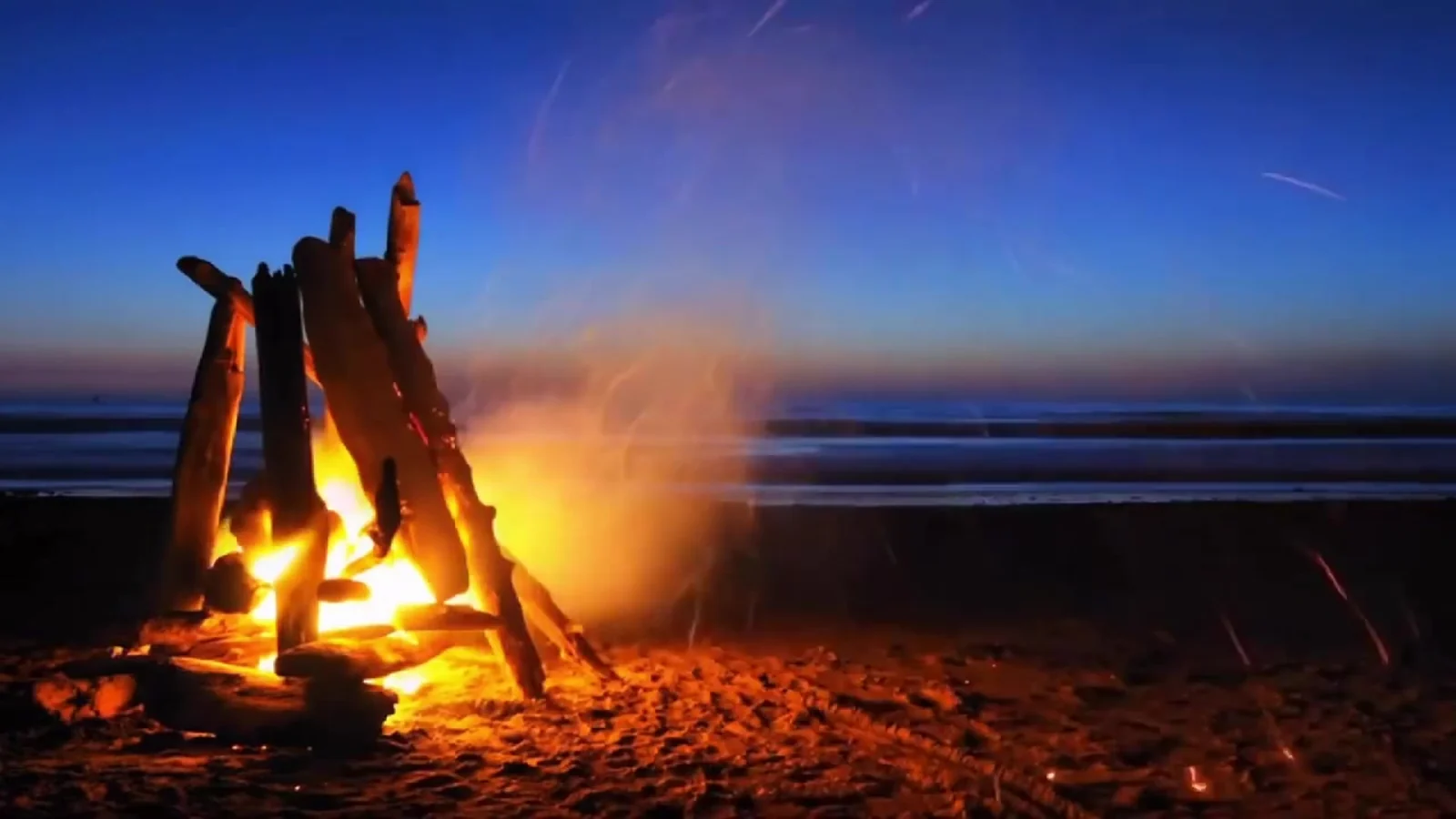 Fotograma del videoclip de «La playa del Mal Pas»: una hoguera de troncos cruzados arde en la arena de la playa al anochecer, con el humo subiendo hacia un cielo azul y el mar al fondo
