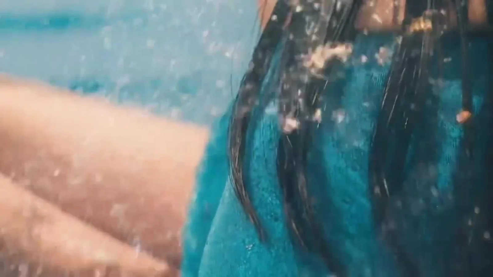 Fotograma del videoclip de «Breeze»: una melena negra mojada cae sobre una tela azul rodeada de gotas de agua, con la arena desenfocada al fondo