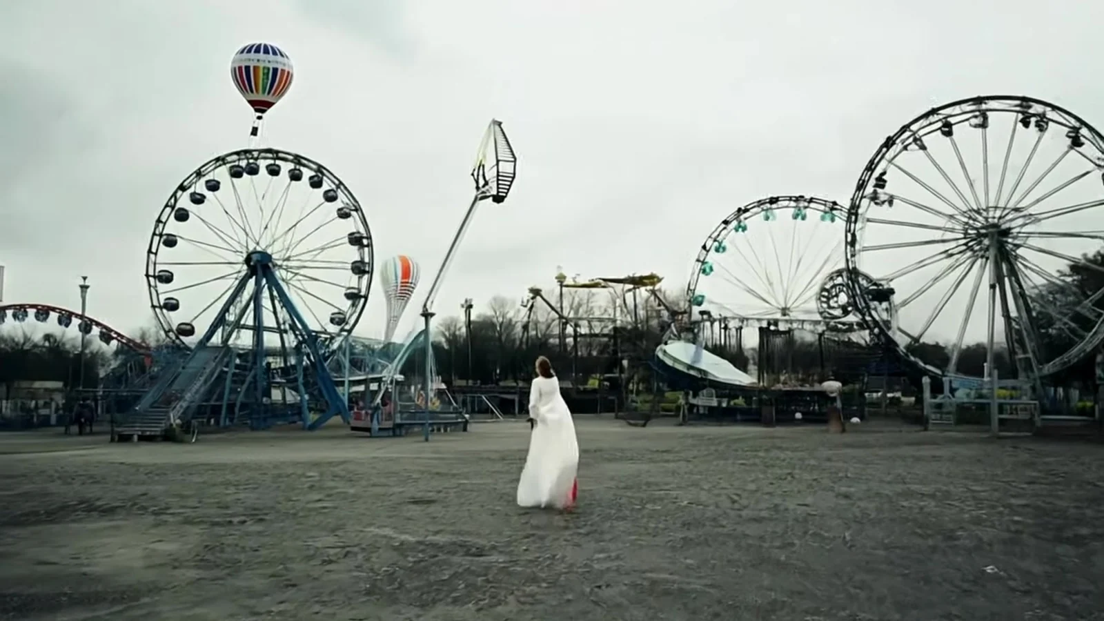 Fotograma del videoclip de «La perspectiva»: tres norias blancas y una montaña rusa en una feria vacía bajo un cielo nublado, con un globo aerostático en lo alto y una figura diminuta de blanco en la explanada