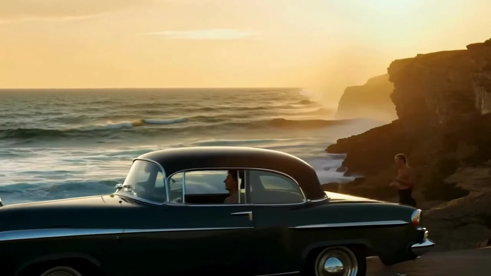 Fotograma del videoclip de «La perspectiva»: un coche antiguo oscuro aparcado en la hierba frente a un acantilado, con el mar dorado por el sol del atardecer