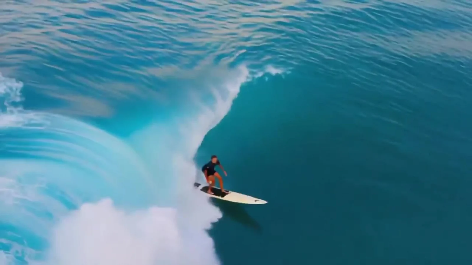 Fotograma del videoclip de «Drift Away»: una ola turquesa gigante se enrosca formando un tubo y un surfista diminuto se desliza dentro sobre su tabla