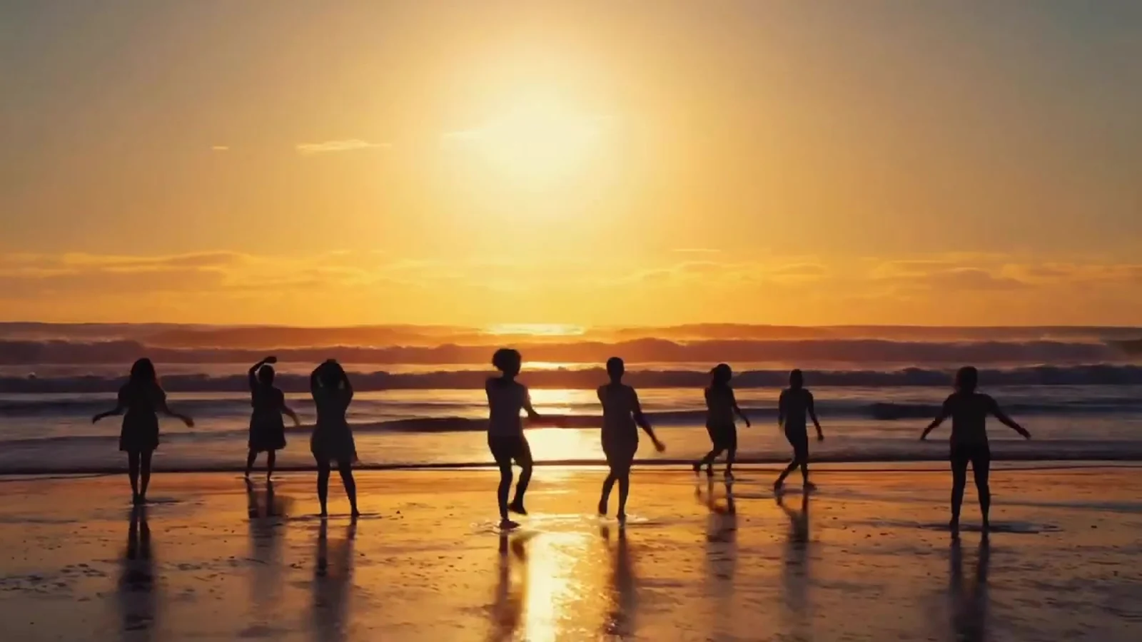 Fotograma del videoclip de «Drift Away»: un grupo de siluetas camina en fila por la orilla frente a un sol enorme que se pone sobre el mar
