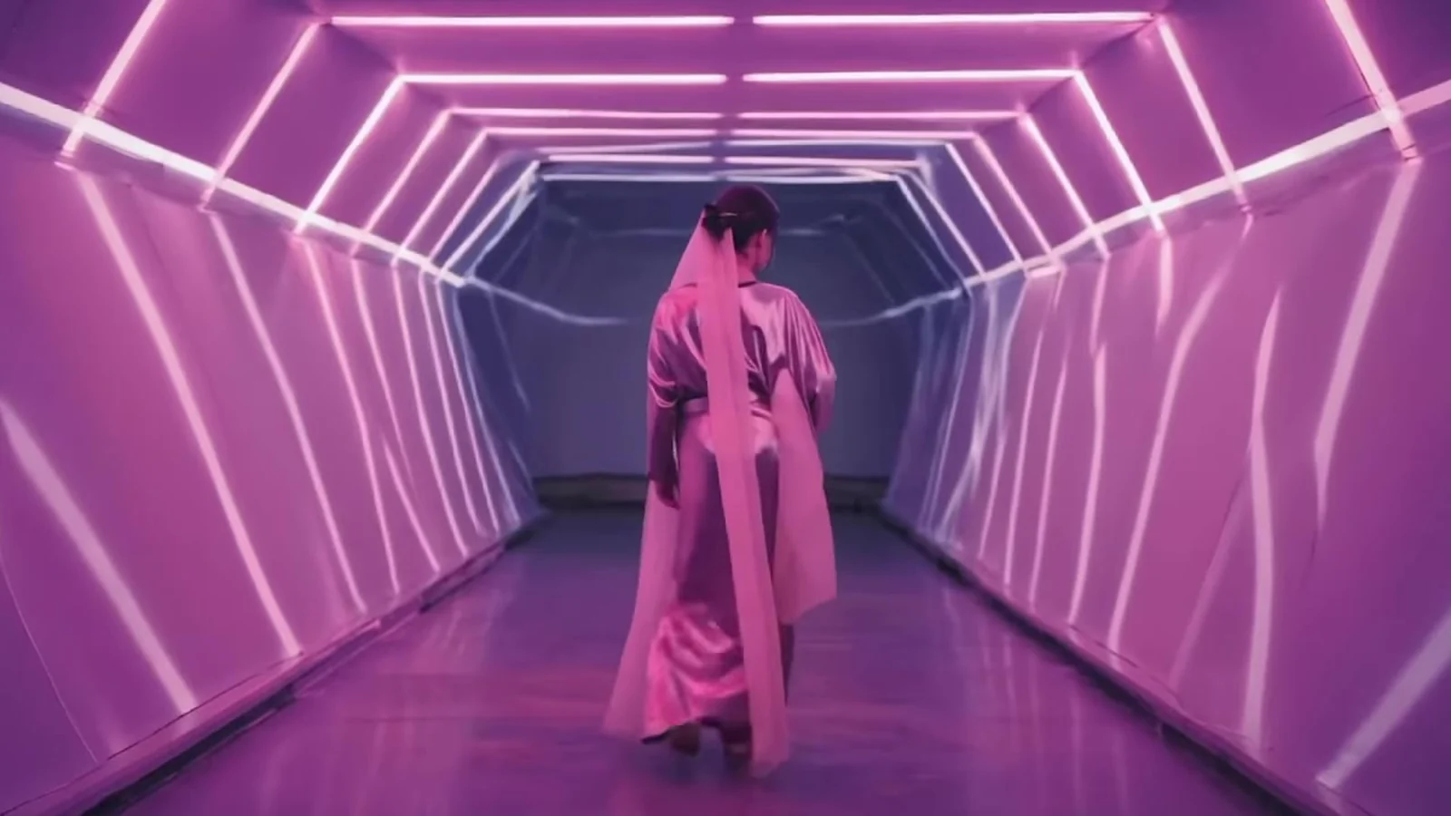 Fotograma del videoclip de «Dubai»: una figura vestida de rosa, de espaldas, camina por un túnel hexagonal de neón rosa y blanco