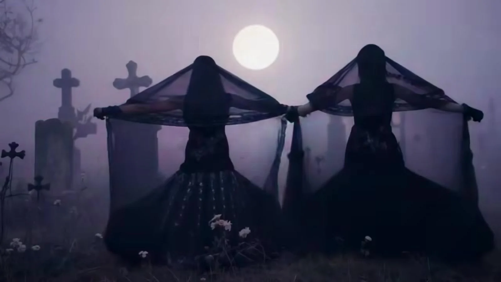 Fotograma del videoclip de «Beat My Post-Traumatic Stress»: dos figuras de negro, de espaldas, abren sus velos oscuros como alas entre cruces de piedra bajo una luna llena
