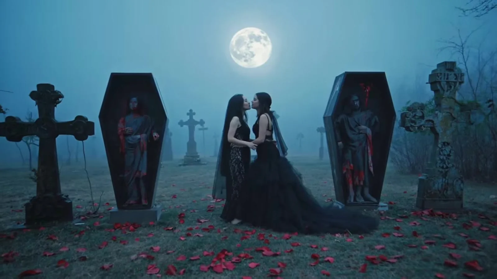 Fotograma del videoclip de «Beat My Post-Traumatic Stress»: dos ataúdes decorados de pie entre lápidas, una alfombra de pétalos rojos y un abrazo bajo la luna llena
