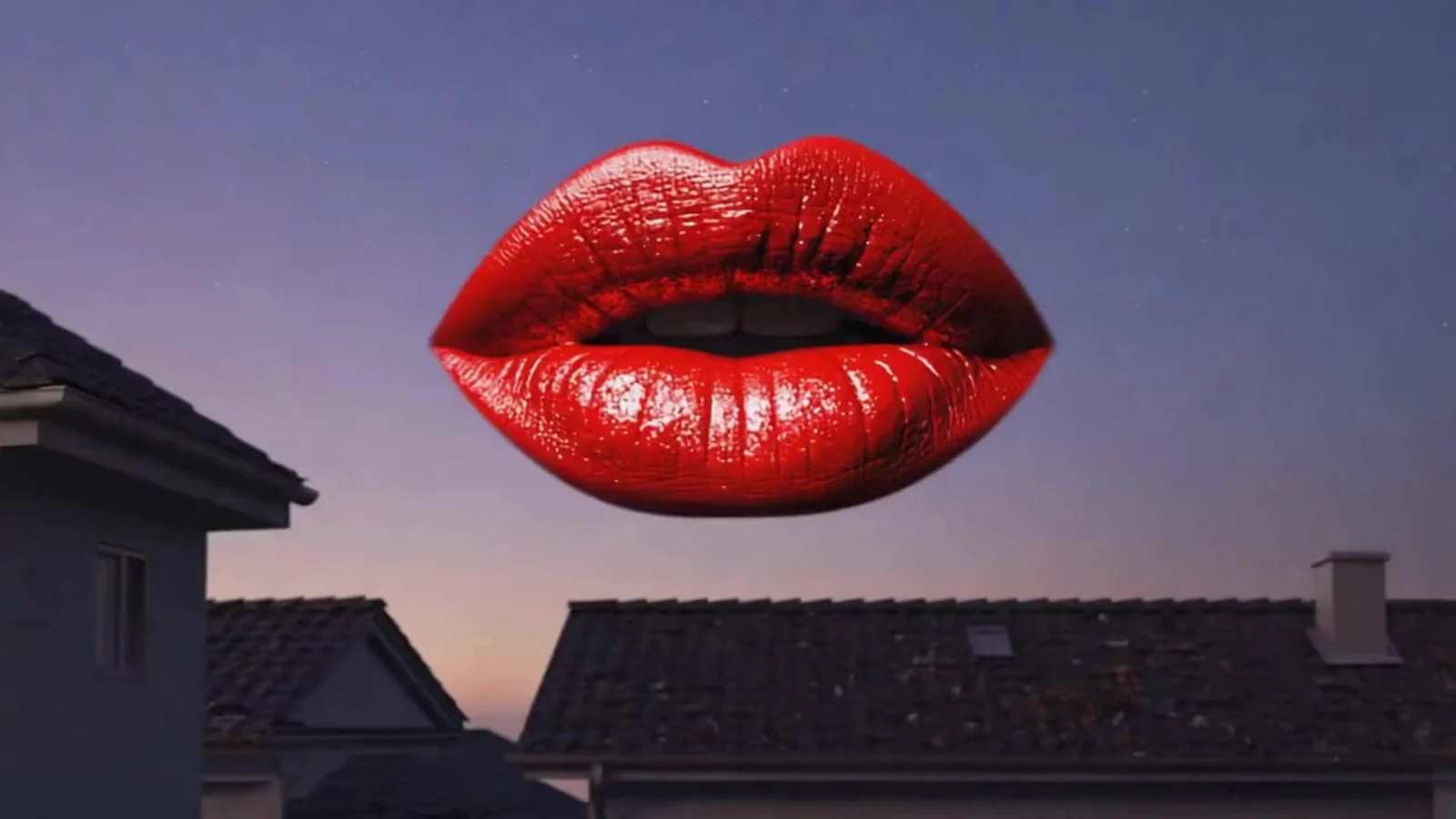 Fotograma del videoclip de «La Boca»: unos labios rojos enormes y brillantes flotan sobre tejados de teja y una chimenea a la hora azul