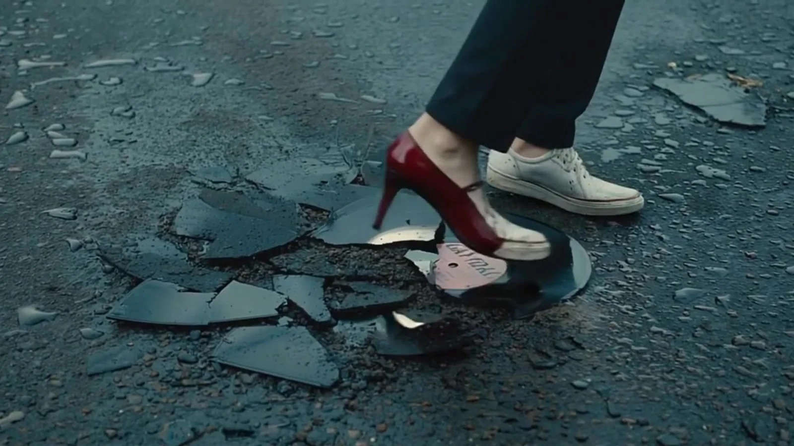 Fotograma del videoclip de «La Boca»: un zapato de tacón rojo y una zapatilla blanca pisan trozos de vinilo roto sobre el asfalto gris
