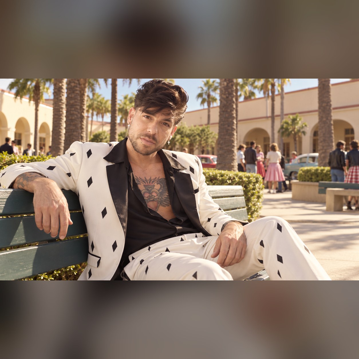 Mike Sain't Louis con traje blanco estampado con rombos negros recostado en un banco de una plaza con palmeras