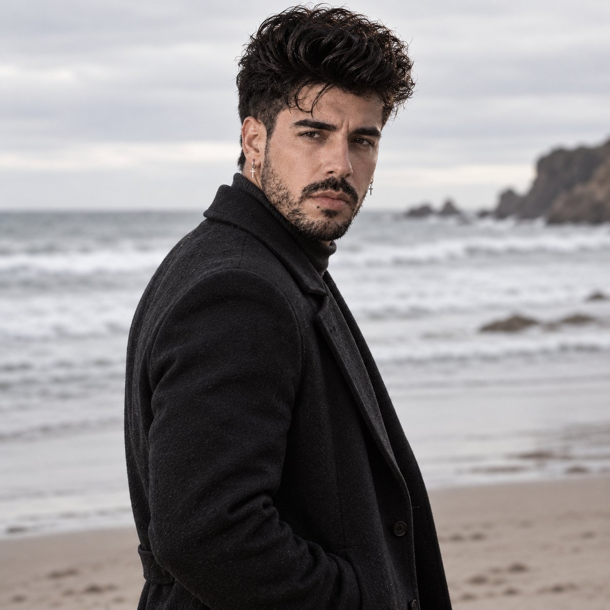 Mike Sain't Louis, hombre de pelo negro rizado y barba, con abrigo de paño negro y suéter de cuello alto, mira por encima del hombro hacia la cámara en una playa de arena con olas grises y rocas oscuras al fondo bajo un cielo nublado.